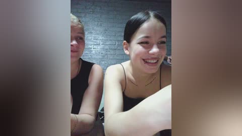 Video of two smiling women with fair skin, one with short blonde hair in a black top, the other with dark hair in a black top, in a dimly lit room with a brick wall background.