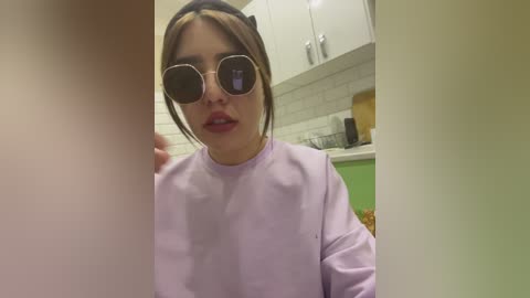 Video of a woman with brown hair, wearing oversized black sunglasses and a light purple top, standing in a modern kitchen with white cabinets and green accents.