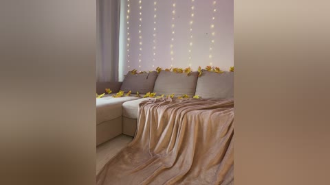 Video of a cozy bedroom with a beige sectional couch, covered in a light brown blanket, adorned with yellow flowers. The wall behind features fairy lights and more flowers, creating a warm, inviting atmosphere.