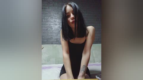 Video of a slender woman with pale skin, long black hair, wearing a black strapless dress, sitting on a bed with a grey brick wall background.