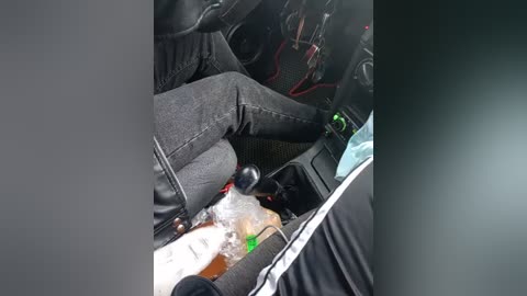 Video of a person's legs and feet in a car, wearing dark jeans, with a beer bottle on the floor, steering wheel visible, red wires in background, blurry interior.