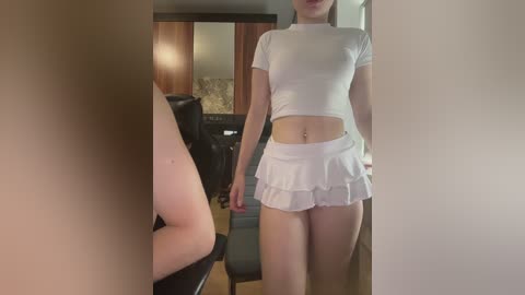 A video of a slim, light-skinned woman in a tight, white crop top and short, ruffled skirt, standing in a dimly lit room with a mirror reflecting a wooden cabinet and black chair.