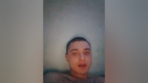 A blurred video of a young man with short dark hair, wearing a gray shirt, against a plain, light-colored wall. His expression is neutral, and the image has a grainy texture.