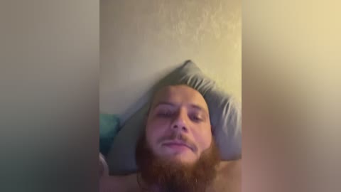 A blurry video of a bearded man with closed eyes, lying on a gray pillow against a beige wall, with a blurred background.