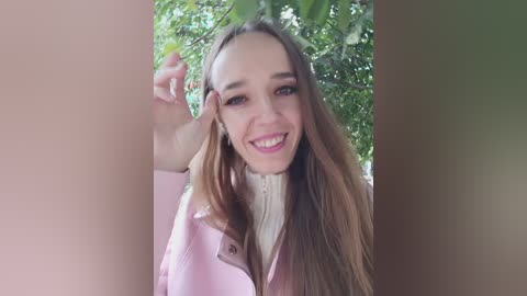 Video of a smiling young Caucasian woman with long brown hair, wearing a pink jacket, standing outdoors among lush green foliage.