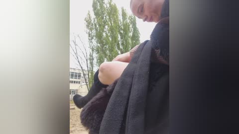 A video of a bald woman with fair skin, wearing black knitwear and dark socks, sitting in a doorway. Trees and a building are visible in the background.