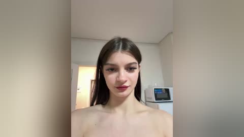 Video of a young woman with fair skin and straight brown hair, standing nude in a modern, minimalist room with beige walls and white furniture.