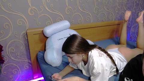 Video of a young girl with long brown hair lying on a blue bed with a plush dog pillow. She's wearing a white t-shirt and white panties. The background features a patterned wallpaper and purple lighting.