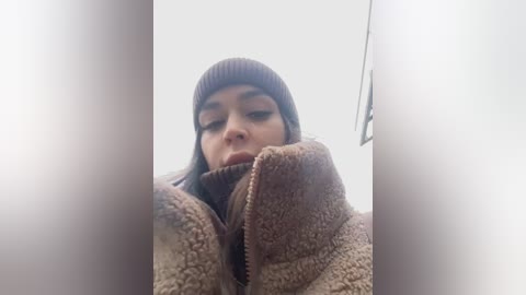 A video of a young woman with long, dark hair, wearing a brown beanie and a textured brown jacket, her face partially obscured by her coat's zipper. The background is blurred and white.