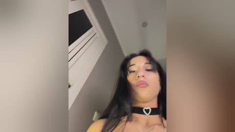 A video of a young woman with long black hair, wearing a black choker with a heart pendant, making a kissing face, in a modern, dimly lit room with white walls and a white ceiling.