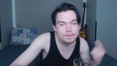 Video of a young man with short dark hair, wearing a black tank top, holding a glass in a dimly lit room with a guitar and a patchwork pillow.