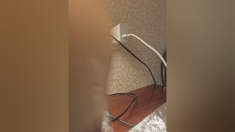 A video of a white electrical outlet with a white extension cord and black wire plugged in, resting on a wooden surface against a wallpapered beige wall with a floral pattern.
