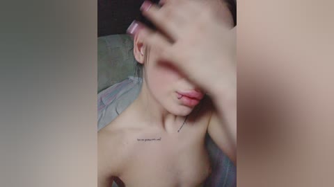 A video of a topless woman with light skin, wearing pastel pink hair, partially covering her face with her hand. She has a small tattoo below her collarbone. The background is a bed with a pastel-colored blanket.