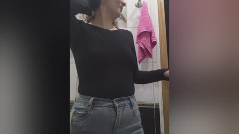 Video of a woman with light skin, wearing a black long-sleeve top and light blue denim shorts, standing in a bathroom with a pink towel hanging on a wooden door.