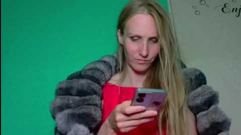 Video of a blonde woman with straight hair wearing a red top and a grey faux fur jacket, looking down at a smartphone with a purple case.