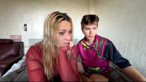 Video of a blonde woman in a red shirt, leaning on a bed, and a young man in a pink and green sports jersey, looking concerned, in a dimly lit room with beige walls.