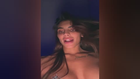 Video of a topless, light-skinned woman with long brown hair, smiling, with a tattoo on her collarbone, against a dark, blurry background.