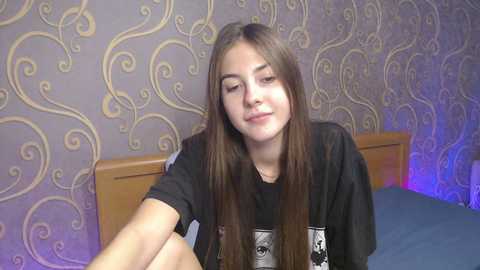 Video of a young woman with long brown hair, wearing a black graphic t-shirt, sitting on a bed with blue sheets. Wallpaper in the background features swirling patterns in muted colors.