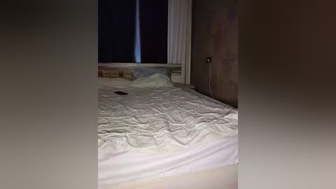 Video of a dimly lit, messy bedroom with a rumpled white bedspread, a black phone on the mattress, and a window partially covered by dark curtains. The room has a muted, earthy color palette.