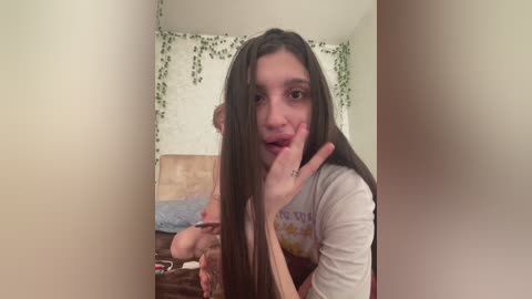 A young woman with long, straight brown hair, wearing a white shirt, sits on a bed with a beige headboard, looking at the camera. The room has green ivy on the walls.