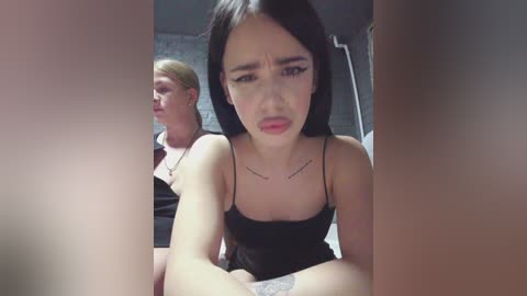 A video featuring two young women in a dimly lit room. The foreground shows a dark-haired woman with a distressed expression, wearing a black spaghetti-strap top. A blonde woman with a neutral expression is behind her.