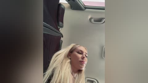 Video of a blonde woman with long hair, wearing makeup, sitting in a car. The background shows a grey car interior with a sunroof and beige seats.