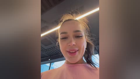 Video of a young Asian woman with light skin, wearing a pink shirt, smiling, with hair tied back, in a modern, well-lit interior with a metal ceiling.
