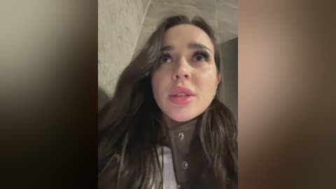 Video of a young woman with long dark hair, light skin, and full lips, wearing a black leather jacket, standing against a textured wall in a dimly lit room.