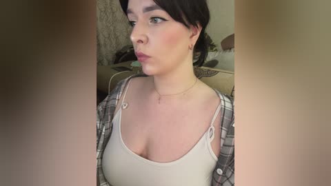 Video of a fair-skinned woman with short black hair, wearing a white tank top, plaid shirt, and gold necklace, looking contemplative in a cozy, dimly lit room with beige furniture.