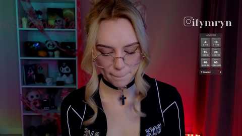 Video of a blonde woman with glasses and a black choker, wearing a black jacket, standing in a room with a red light, shelves with stuffed toys, and a \"Lymmr\" watermark.