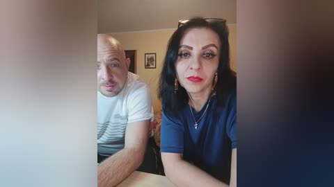 Video of a bald, white man with a mustache in a white striped shirt and a woman with black hair, red lipstick, and a blue top, sitting closely at a table, blurred background.