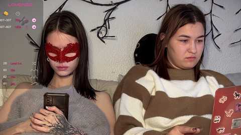 Video of two young women in a living room, one wearing a red Venetian mask, the other a striped sweater, both focused on smartphones. Background shows a white wall with abstract black art.