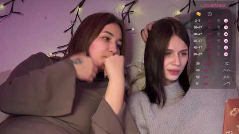 Video of two young women with straight, dark hair, one kissing the other's shoulder, wearing casual clothes in a dimly lit room with string lights.