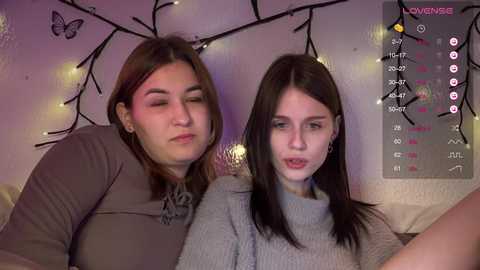 Video of two young women with fair skin, one in a grey sweater, the other in a beige top, sitting closely on a couch. Background features a white wall with black tree branches and fairy lights.