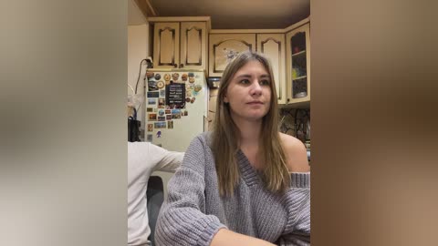 Video of a young woman with long, straight brown hair, wearing a gray, off-shoulder knitted sweater, seated in a cozy, rustic kitchen with wooden cabinets, a collage of photos on the fridge, and a cluttered countertop.