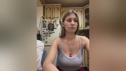 Video of a young Caucasian woman with straight, shoulder-length blonde hair, wearing a grey tank top and red skirt, sitting in a cluttered kitchen with beige cabinets, a fridge adorned with photos, and a cluttered countertop.
