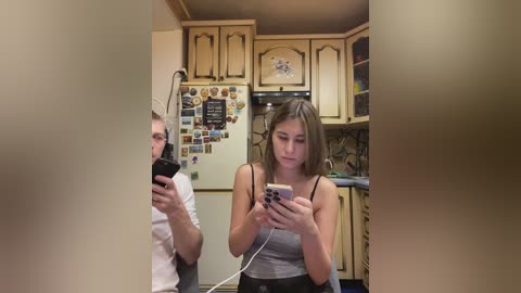Video of a young woman with light skin and brown hair, wearing a grey tank top, standing in a kitchen with wooden cabinets, a fridge adorned with magnets, and a man in the background.