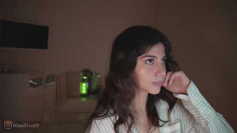 Video of a young woman with long, wavy black hair, light skin, and brown eyes, wearing a white ribbed sweater, looking contemplative in a dimly lit room with a muted color palette and a green-lit water bottle in the background.