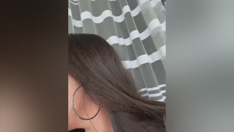Video of a woman with long, straight, dark brown hair, wearing large silver hoop earrings, standing in a bathroom with a white and gray striped shower curtain.