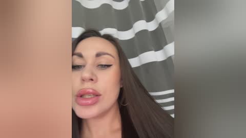 Video of a young woman with fair skin, long brown hair, and light pink lipstick, wearing a black top. She has a pouty expression and is partially obscured by a white and gray striped shower curtain.