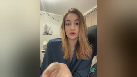 A video of a young woman with long, straight blonde hair and light skin, wearing a blue shirt, sitting in a dimly lit room with a blurred background.