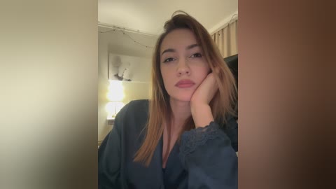 Video of a young woman with light skin and straight, shoulder-length brown hair, wearing a dark blue robe, resting her chin on her hand in a dimly lit room.