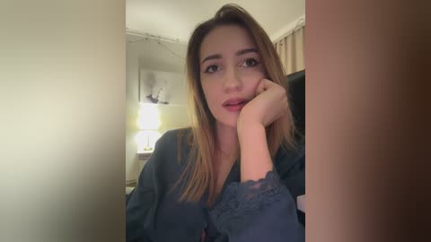 Video of a young woman with light skin and straight, shoulder-length brown hair, wearing a blue lace robe, leaning on a table with a lamp in the background.