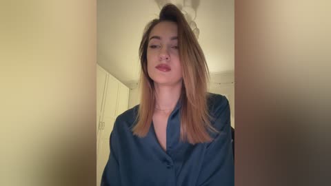 Video of a young woman with fair skin, shoulder-length blonde hair, and red lipstick, wearing a loose-fitting dark blue shirt, indoors against a cream-colored wall.