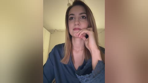 A video of a young woman with light skin, straight brown hair, and a slight smile, wearing a dark blue sweater, posing indoors against beige walls.