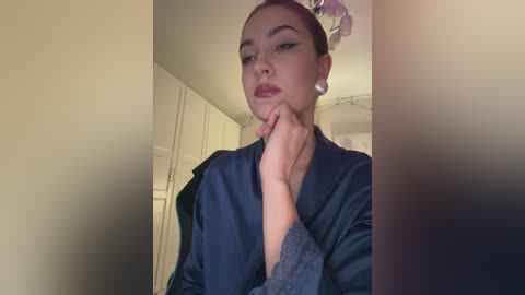 Video of a light-skinned woman with dark hair styled in a bun, wearing a navy satin robe and pearl earrings, in a dimly lit room with cream walls and a floral ceiling fixture.