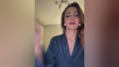 Video of a young woman with light skin, brown hair, and a V-neck blue robe, standing in a dimly lit room with beige walls and a ceiling light fixture.