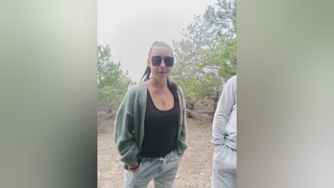 Video of a woman with a medium build, wearing a green jacket, black tank top, and gray sweatpants, standing outdoors near pine trees under a cloudy sky.