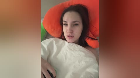 Video of a young woman with fair skin and dark hair, lying on a bed with a white pillow and a bright orange cushion, looking tired and slightly pensive.