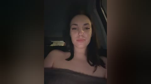 A dimly lit video of a young woman with long, dark hair, wearing a black strapless top, seated in a car.